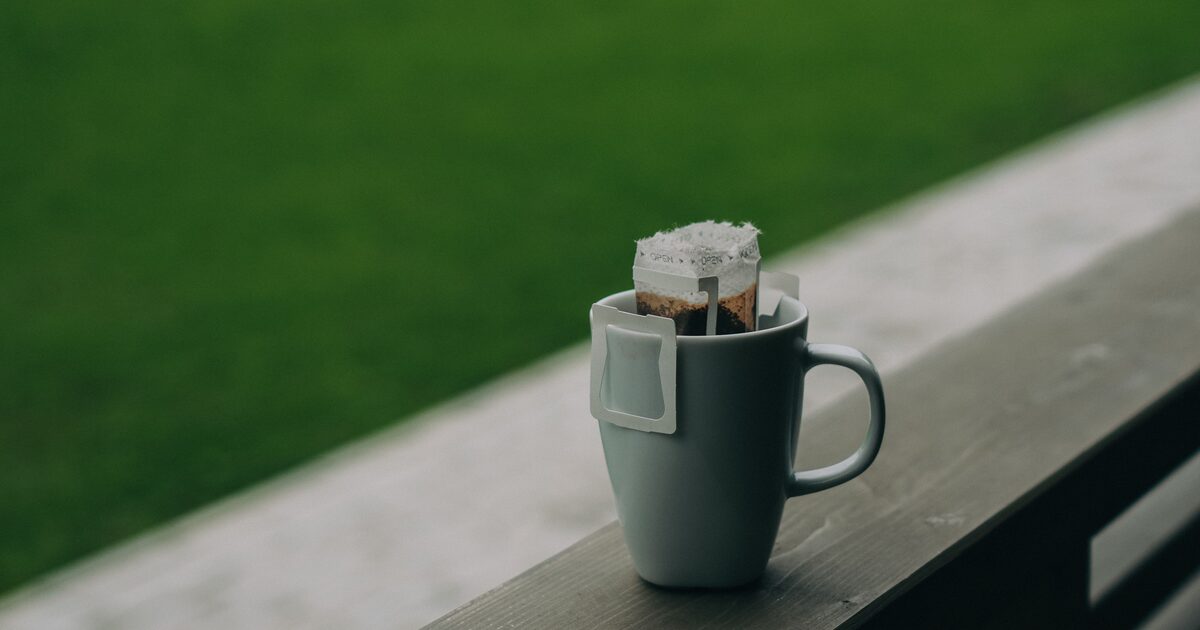 Caneca com sachê de drip coffee encaixado na borda, apoiada num corrimão de madeira ao ar livre