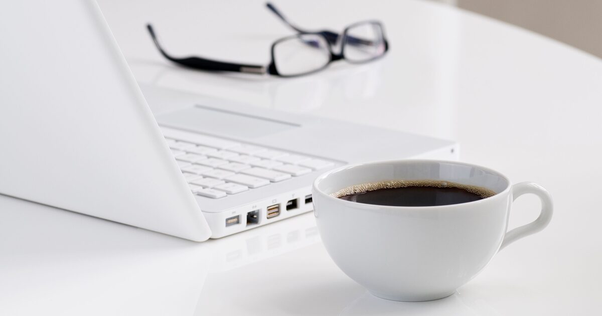 Xícara de café preto ao lado de um laptop e óculos numa mesa branca de escritório